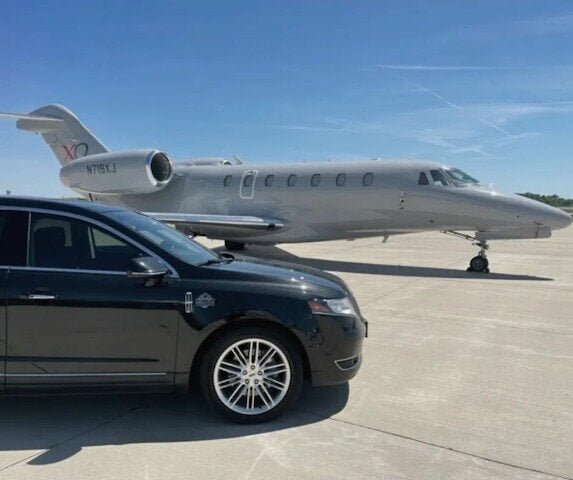 Black executive sedan parked beside a private aircraft on the ramp