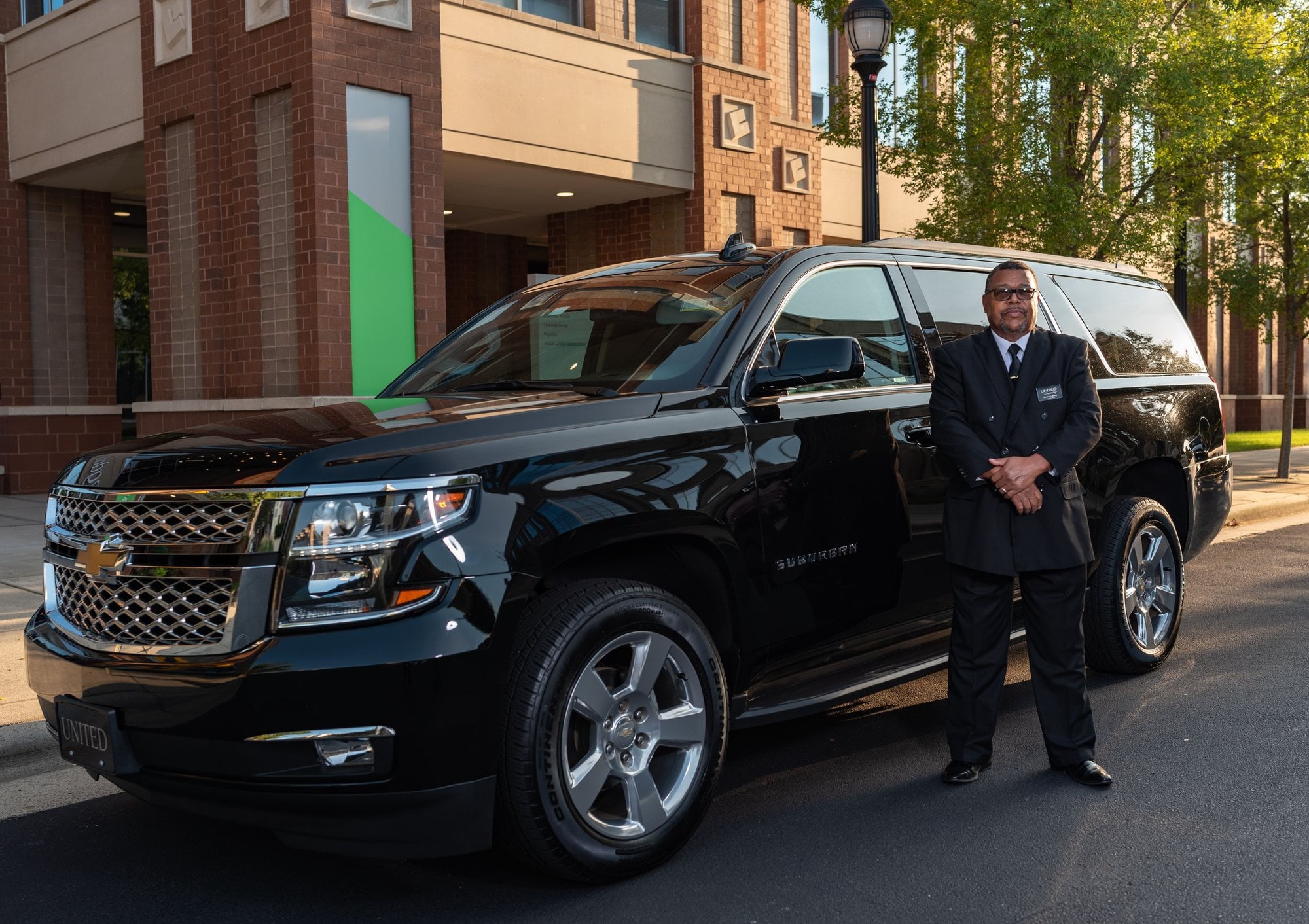A chauffeur in a dark suit stands beside a black full-size SUV outside a brick building.