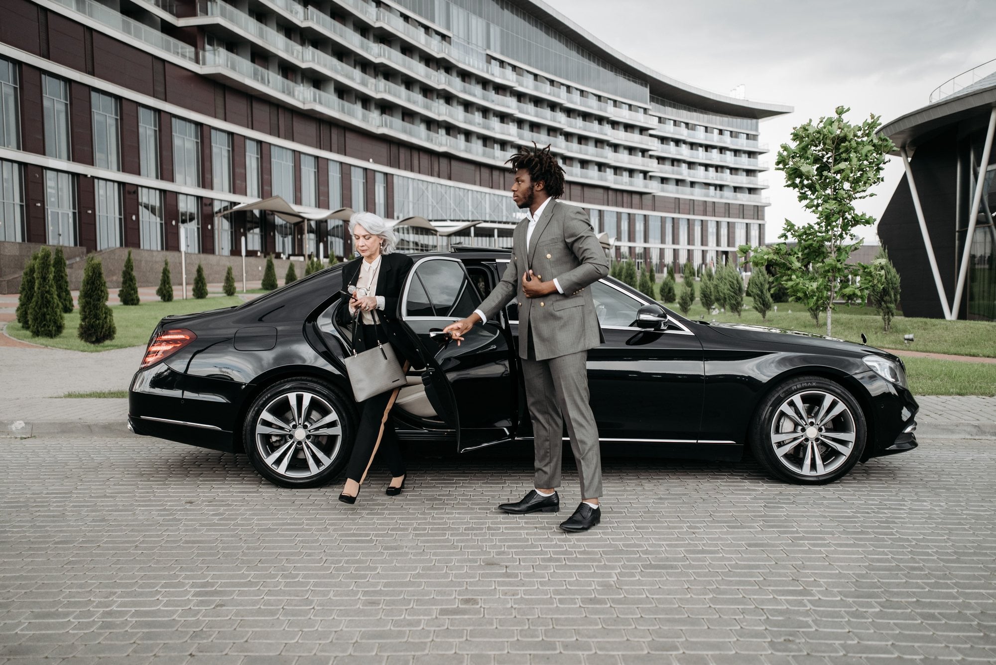 A chauffeur holds the rear door as an executive steps out of a black luxury sedan outside a modern building.