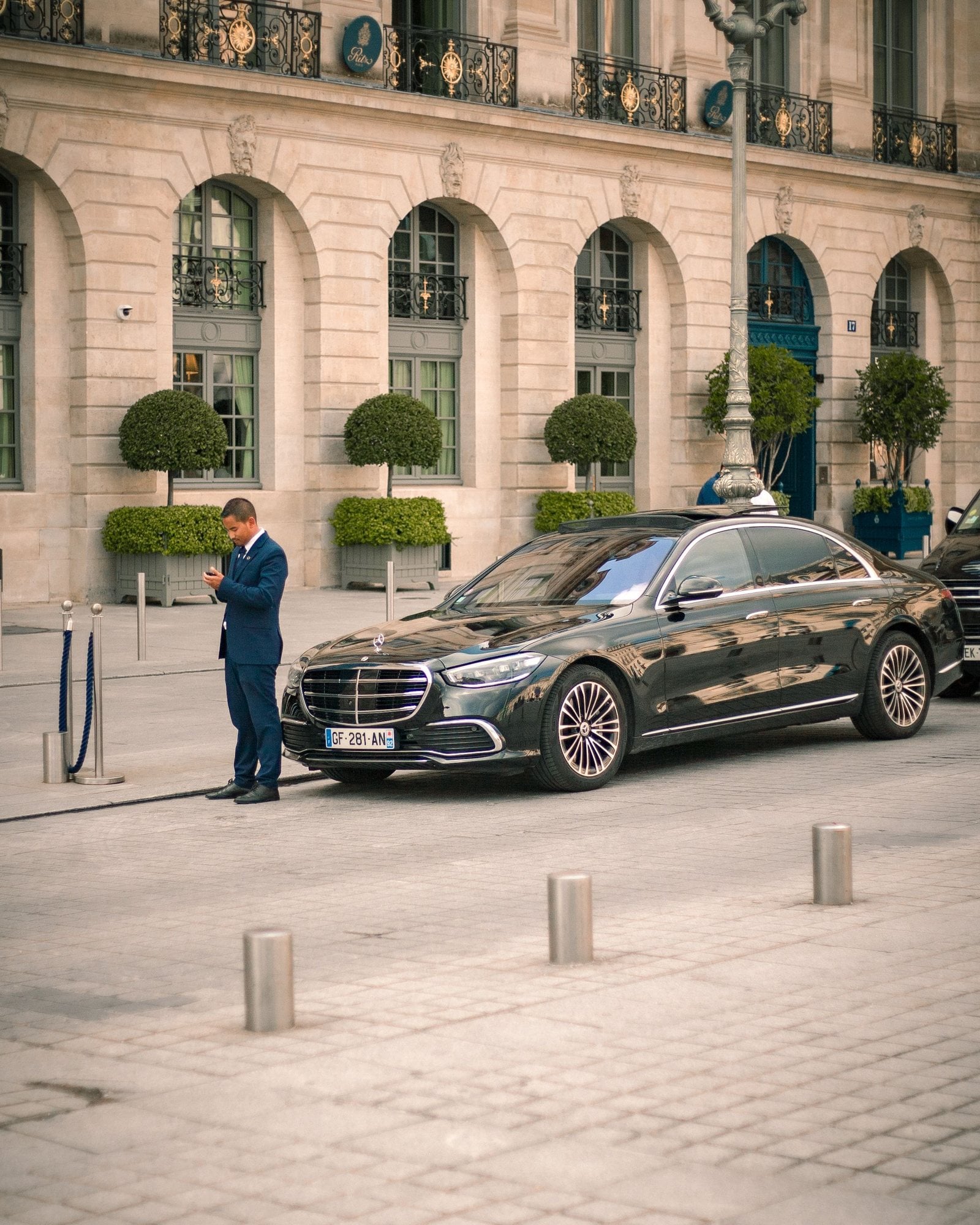 A man in a black suit stands beside a dark luxury car on a city street.
