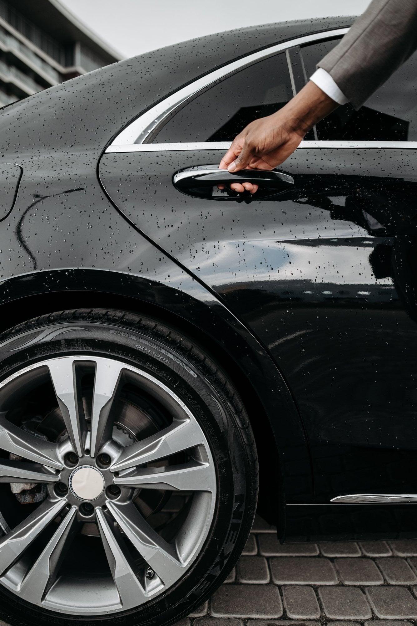A hand opens the rear door of a black car covered in raindrops.
