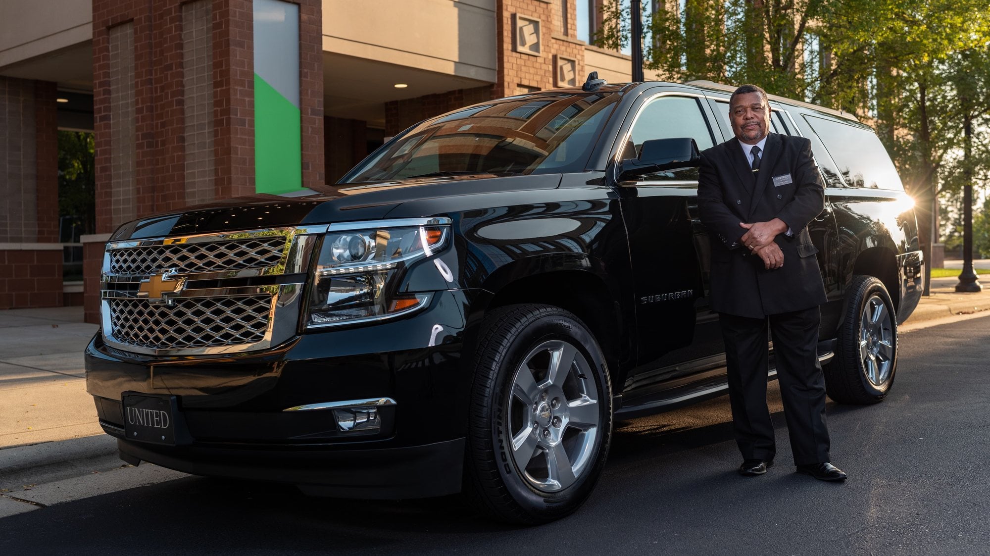 A uniformed chauffeur standing at the passenger door of a black full-size SUV on a city street