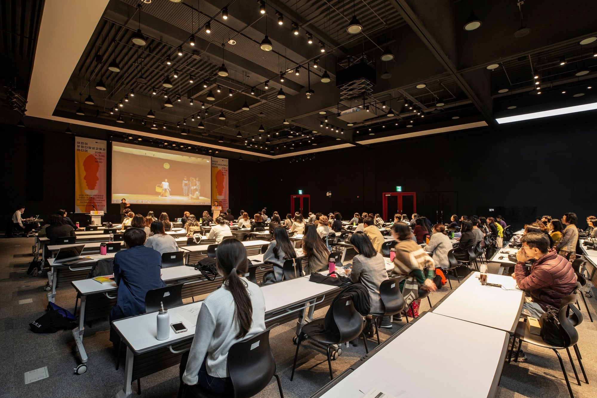 Conference attendees sit at rows of tables facing a presentation screen in a large meeting hall.