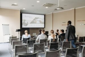 Attendees sit in rows facing a projection screen during a business presentation in a meeting room.