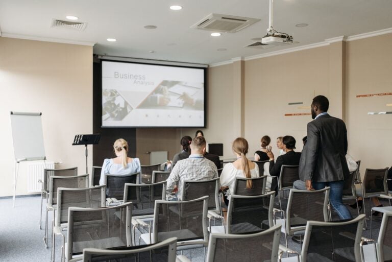 Attendees sit in rows facing a projection screen during a business presentation in a meeting room.