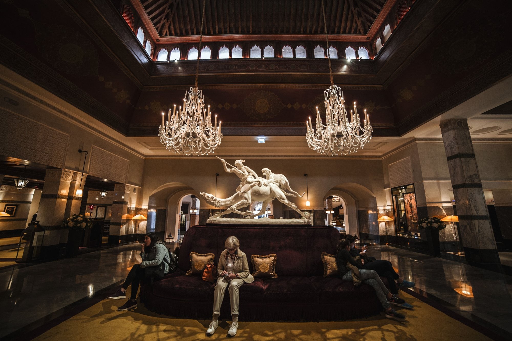 Guests sit beneath chandeliers in the spacious marble lobby of a luxury hotel.