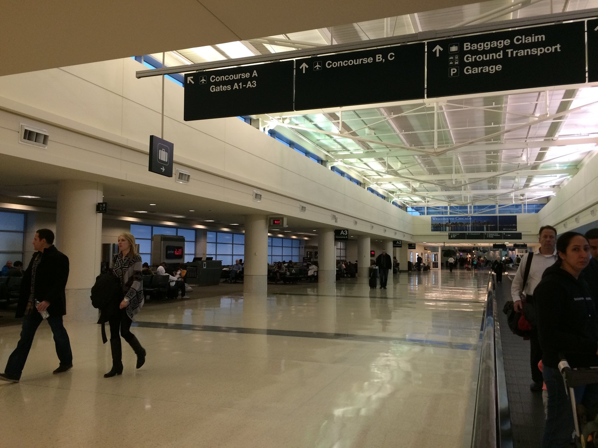 Travelers walking under overhead concourse, baggage claim and garage signs inside Chicago Midway International Airport