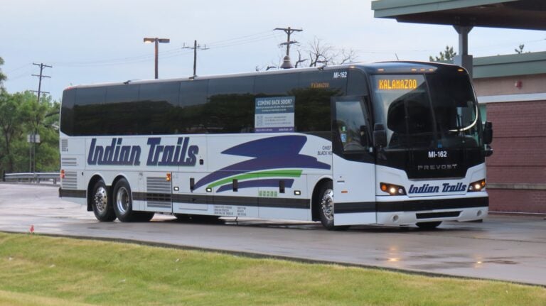 A white Prevost X3-45 motorcoach parked under a covered station canopy with its destination sign lit.