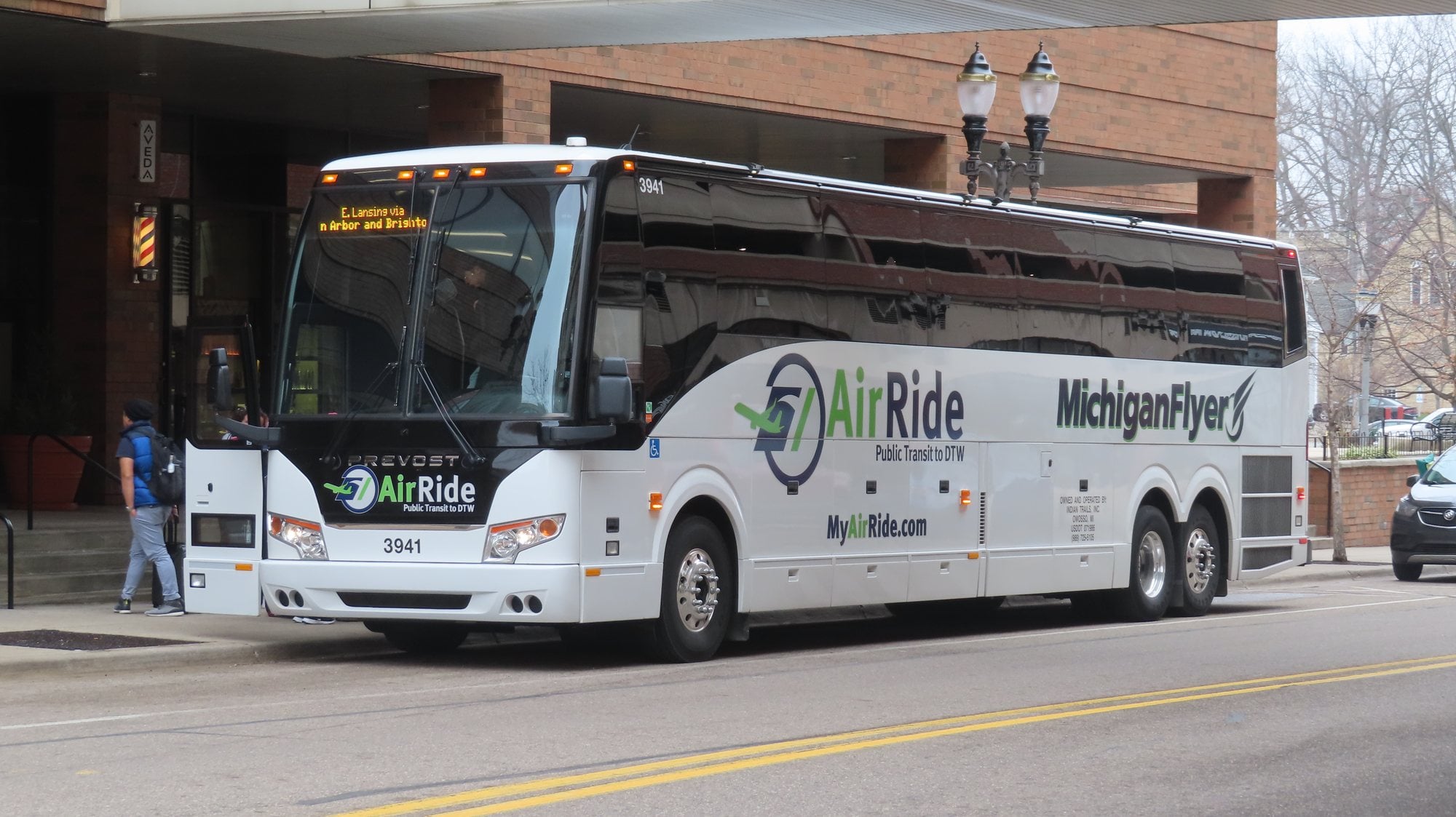 A white motorcoach waits on a downtown street while a passenger with a backpack walks toward the open door.