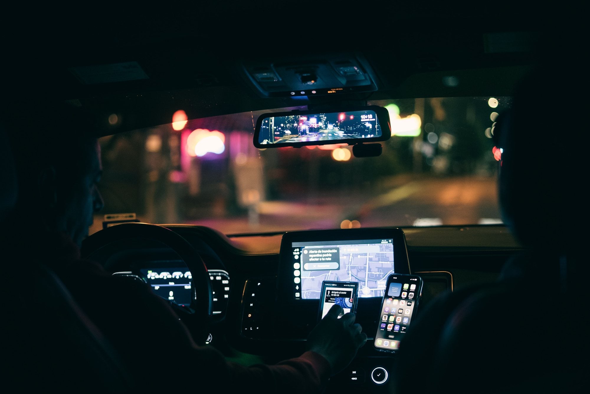 At night, a driver follows navigation on dashboard-mounted smartphones as city lights blur through the windshield.