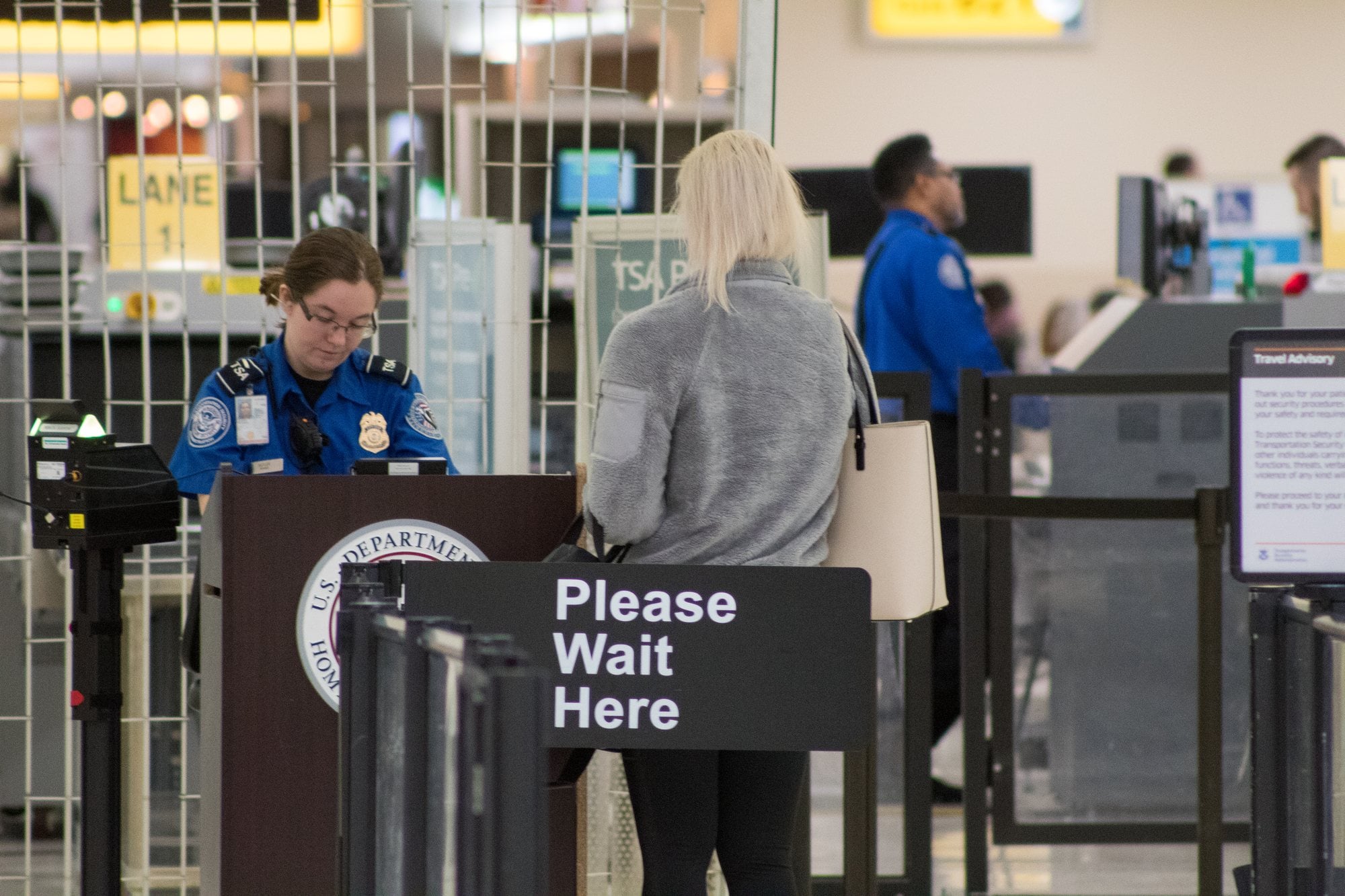 A traveler waiting behind a Please Wait Here sign at a TSA document-check podium at an airport security checkpoint